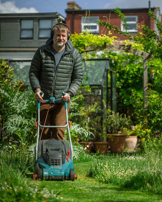 A middle-aged man with short, gray hair and a beard is operating a garden lawn mower on a lush, well-maintained lawn. He is dressed in a black, quilted jacket and brown trousers, wearing grey gloves f